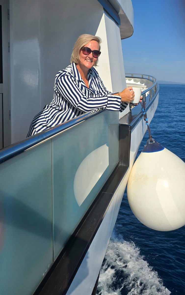 SJ wearing sunglasses and a striped PJs leans on the railing of a luxury cruise boat, holding a cup.