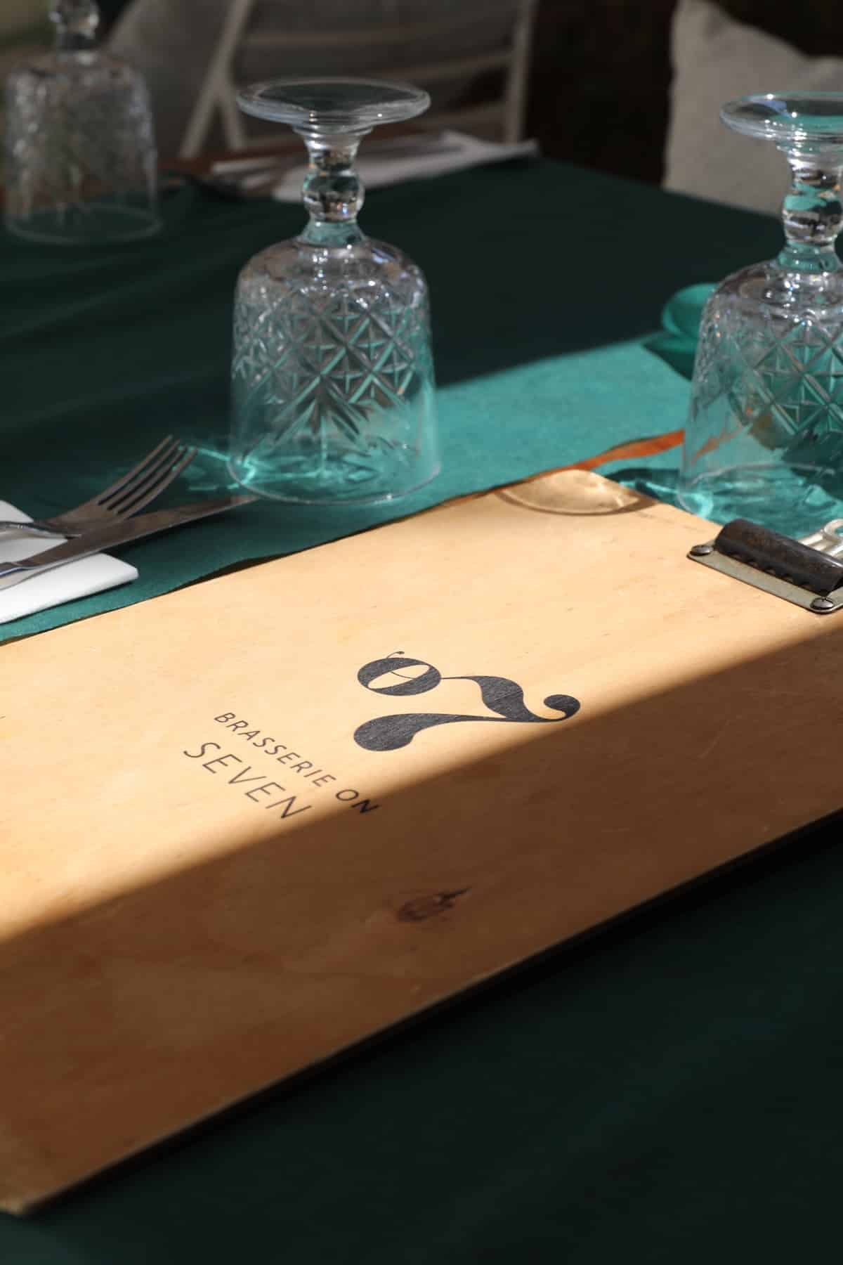 A wooden menu with the text "Brasserie on Seven" lies on a green tablecloth, surrounded by upside-down glasses and cutlery—perfect for a chic hen party in Split, Croatia. B7 Split