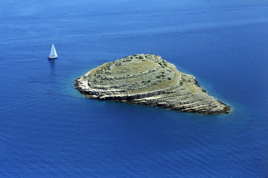 An aerial view of a small, rocky island surrounded by blue water, with a single sailboat nearby—perfect for trips to Kornati National Park.